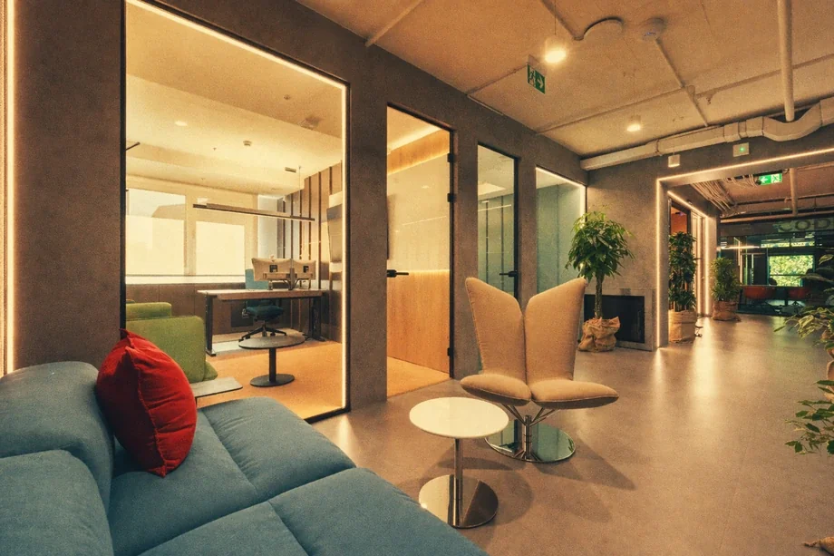 Hallway with blue sofa beside a glass-walled office with orange designer chairs at Coinis HQ