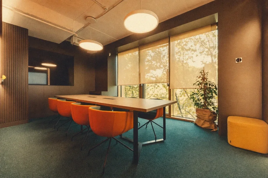 Meeting room with orange swivel chairs around a dark table, sheer blinds, and teal carpet at Coinis HQ